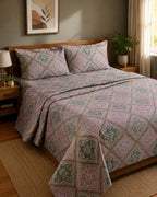 A bed dressed in a patterned quilt and matching pillow shams with a geometric block-print style design in muted purple, sage green, and beige tones, styled in a warm, modern bedroom setting.