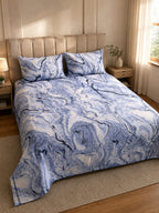 A bed dressed in a blue and white marble-patterned duvet cover and matching pillowcases, styled in a warm, modern bedroom with beige walls, wooden nightstands, and a window with sheer curtains.
