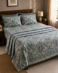 A neatly made bed in a bedroom setting, featuring a light blue and teal patterned quilt, matching pillow shams, and solid teal pillows against a dark wood headboard.