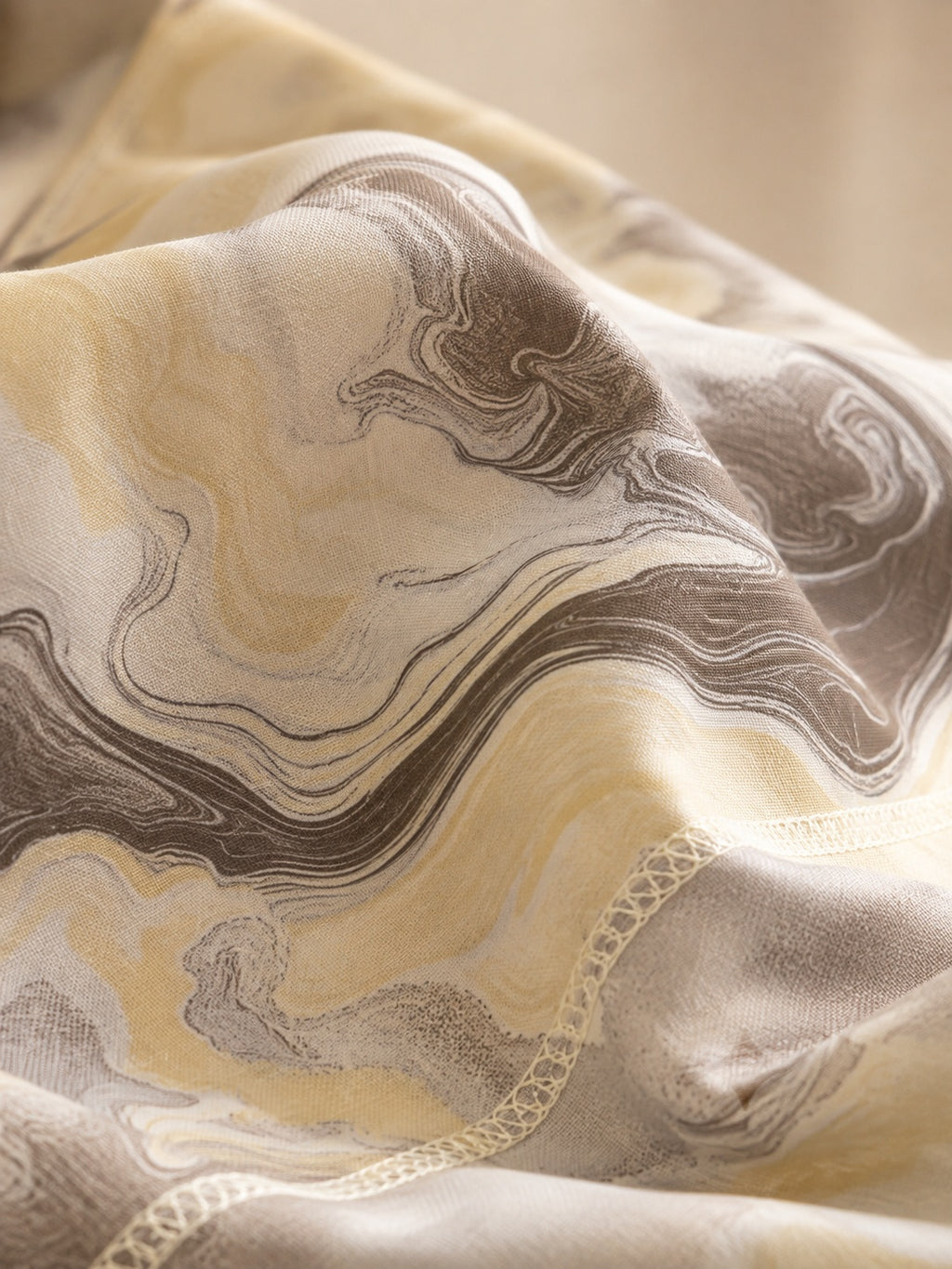A close-up of draped fabric with an abstract marbled pattern in beige, cream, taupe, and gray tones, with a subtle scalloped edge visible.