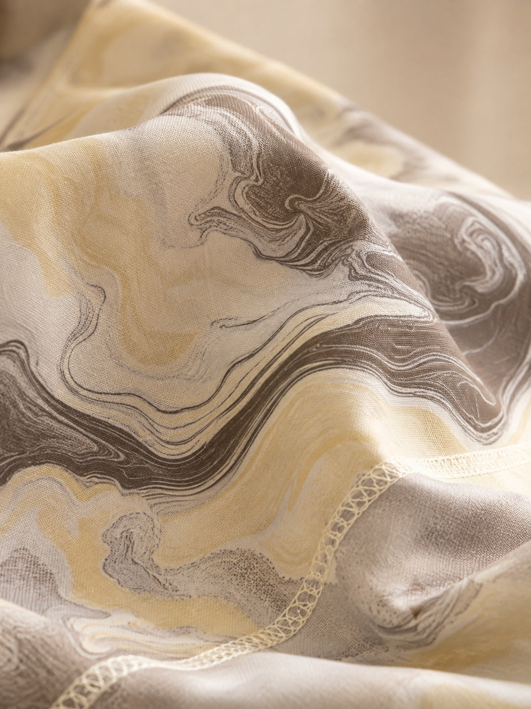 A close-up of draped fabric with an abstract marbled pattern in beige, cream, taupe, and gray tones, with a subtle scalloped edge visible.
