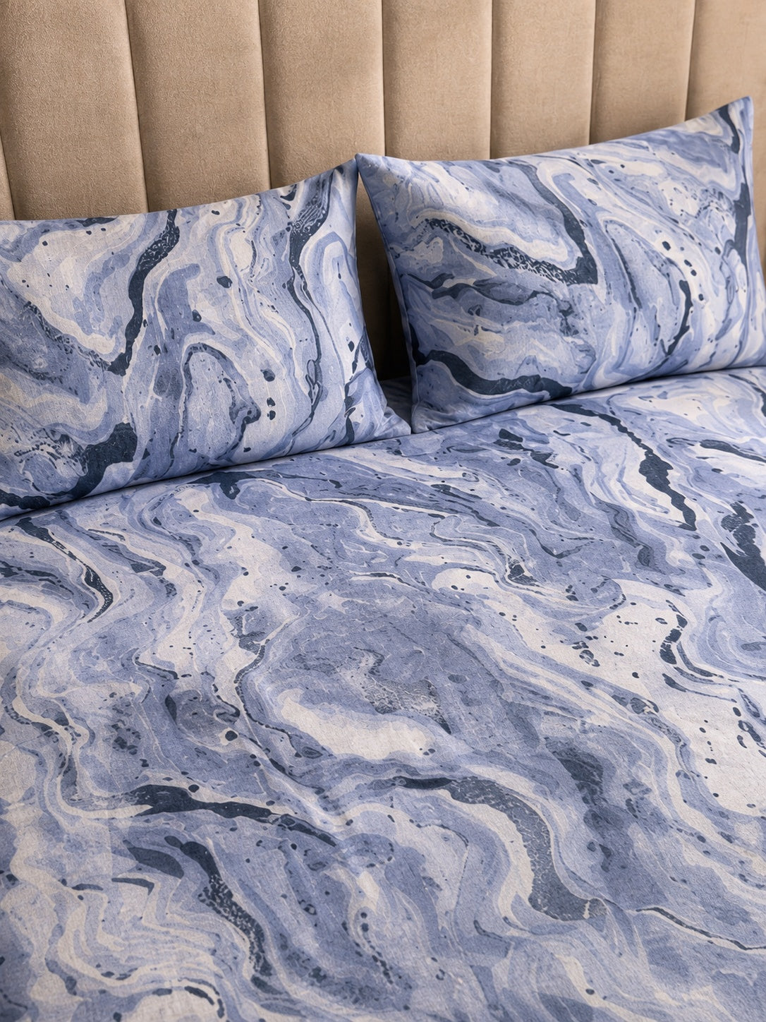 Blue and white marble-patterned bedding set with two matching pillowcases on a bed against a beige tufted headboard.