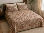 A bed dressed in a beige quilt and matching pillow shams with an ornate damask-style floral pattern in muted red, green, and brown tones, styled in a warm bedroom setting with a wooden headboard, nightstand, plants, and curtains.