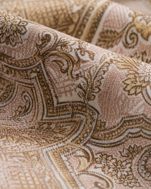 A close-up of draped fabric with an ornate paisley and scroll pattern in warm beige, tan, and brown tones on a light cream background.