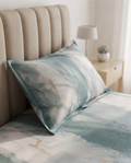 A decorative pillow and matching duvet cover with an abstract watercolor pattern in shades of teal, blue, and cream, styled on a bed with a beige tufted headboard and a bedside table in the background.