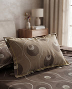 A decorative rectangular pillow with a taupe and brown abstract circular pattern, resting on a matching patterned bedspread in a softly lit bedroom setting.