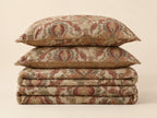 A stack of three decorative pillows and folded matching textiles with an ornate floral tapestry pattern in muted rust, beige, and olive tones, photographed on a plain warm cream background.