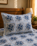 A decorative throw pillow with a blue and gray floral mosaic pattern rests on a bed with matching patterned bedding, set in a warm, sunlit bedroom.