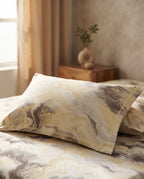 A close-up of a bed in a softly lit bedroom, featuring a pillow and matching bedding with an abstract marbled pattern in cream, beige, gray, and muted gold tones.