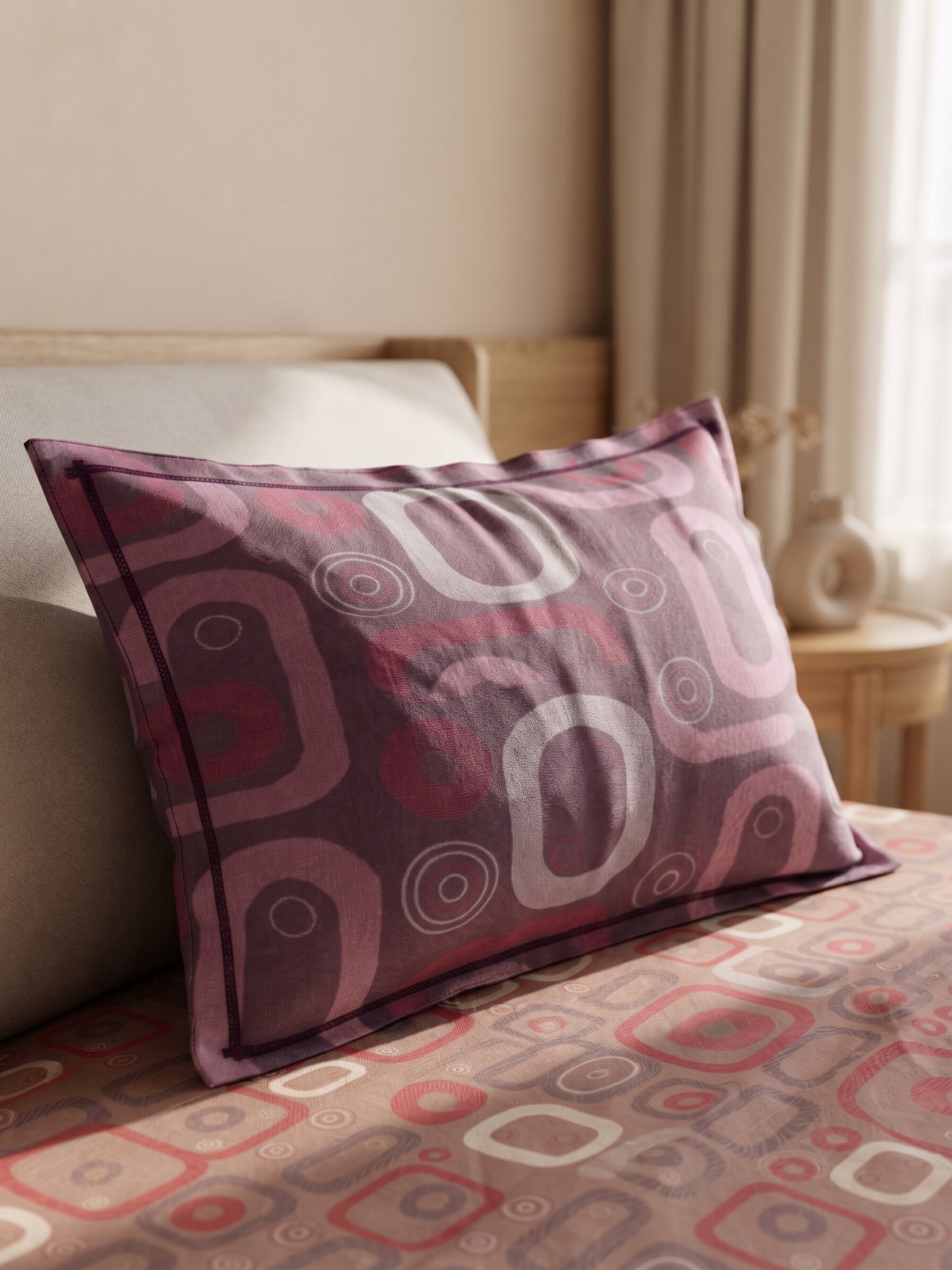 A rectangular decorative pillow with a retro geometric pattern in shades of purple, pink, and white, resting on a bed with a matching patterned sheet.
