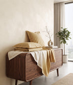 A mid-century modern wooden sideboard cabinet in a warm walnut finish, styled with gold and cream bedding, pillows, a throw blanket, a small vase with branches, and a potted plant in a bright, minimalist room.