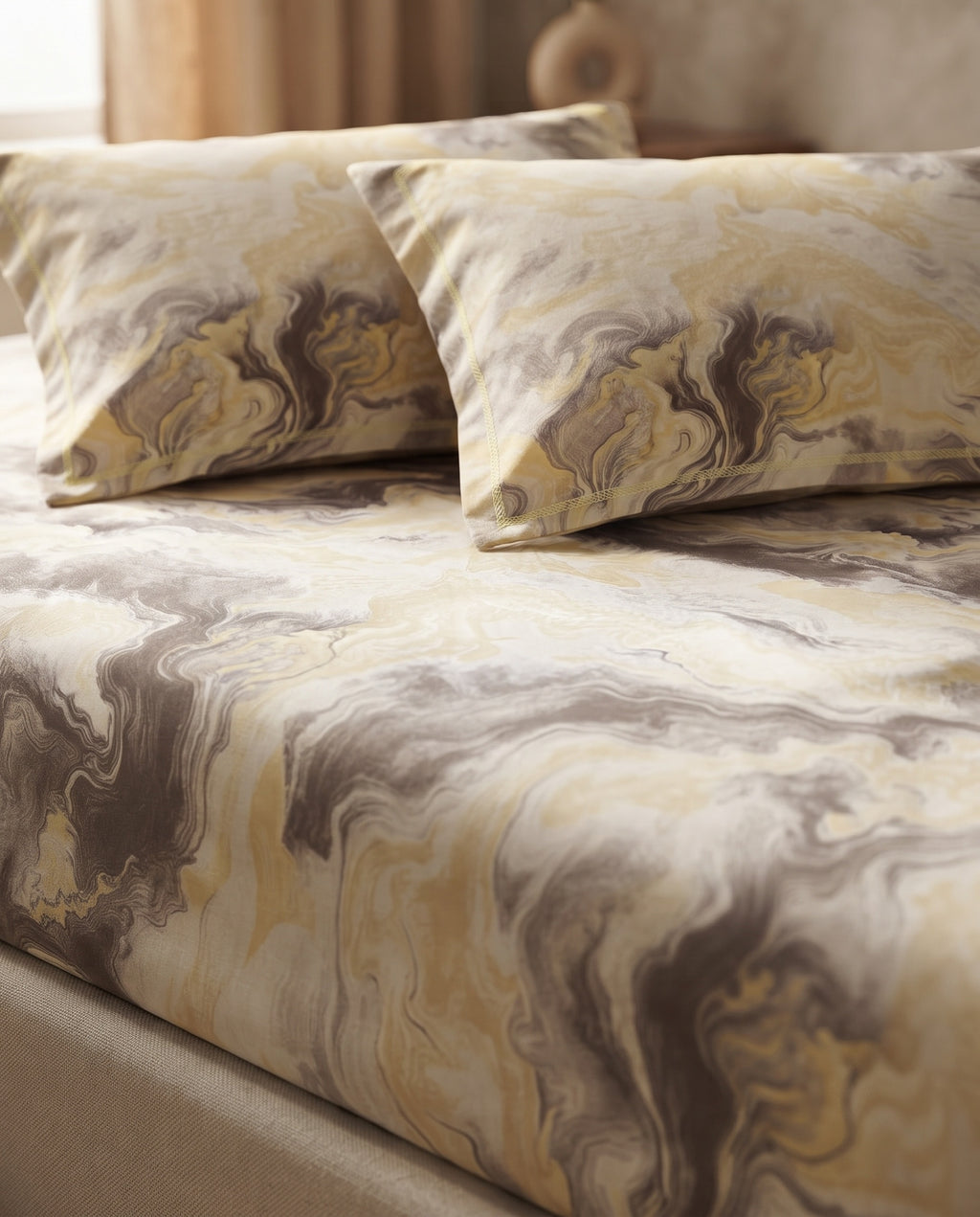 A close-up of a bed dressed in a marbled patterned bedding set with two matching pillowcases. The fabric features an abstract swirl design in cream, beige, mustard yellow, and taupe-brown tones.