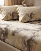 A close-up of a bed dressed in a marbled patterned bedding set with two matching pillowcases. The fabric features an abstract swirl design in cream, beige, mustard yellow, and taupe-brown tones.