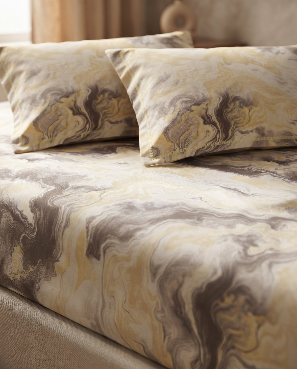 A close-up of a bed dressed in a marbled patterned bedding set with two matching pillowcases. The fabric features an abstract swirl design in cream, beige, mustard yellow, and taupe-brown tones.