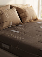 A bed dressed in a brown geometric-patterned sheet and matching pillowcases, photographed in a softly lit bedroom setting.