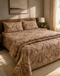 A neatly made bed in a warm, softly lit bedroom featuring a beige quilt and matching pillow shams with an intricate rust-red and olive green paisley pattern. The bed is styled with a dark wood headboard, a bedside lamp, and a neutral-toned wall with a framed painting.
