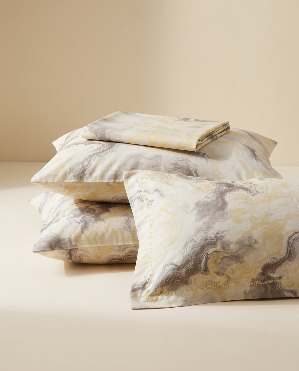 A stack of decorative pillows and a folded matching pillowcase set with an abstract marbled pattern in cream, beige, gray, and muted gold tones, photographed on a warm beige studio background.