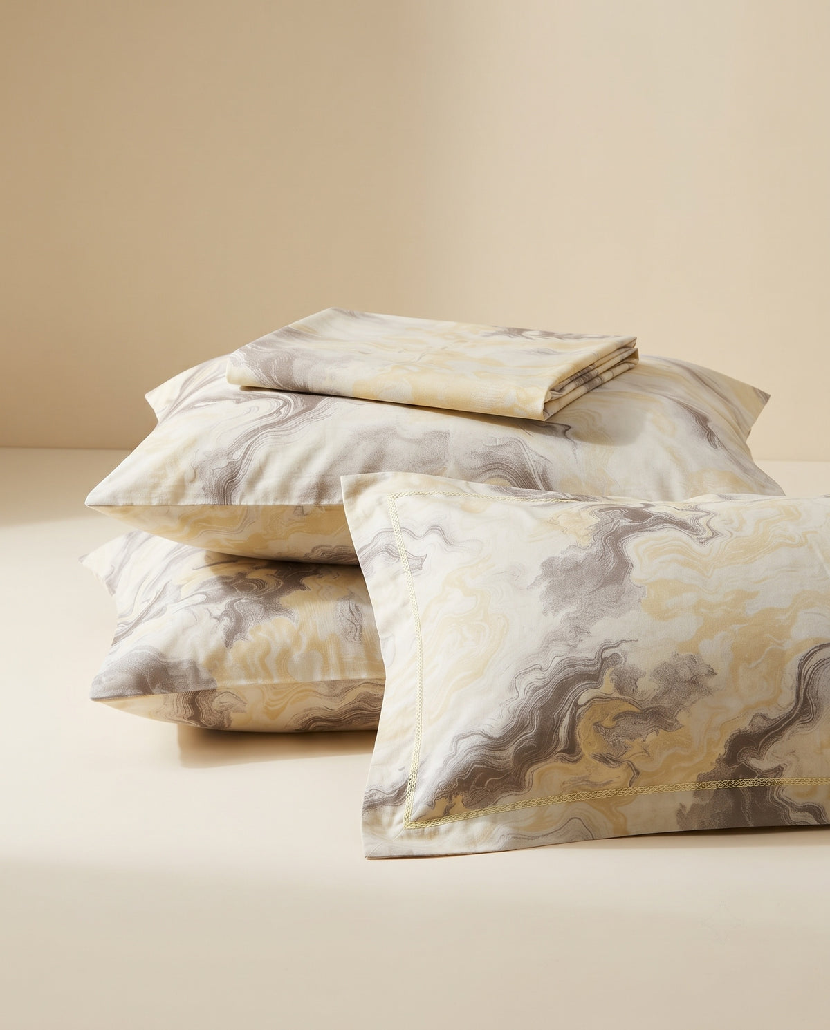 A stack of decorative pillows and a folded matching pillowcase set with an abstract marbled pattern in cream, beige, gray, and muted gold tones, photographed on a warm beige studio background.