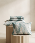 A set of bedding pieces with a teal and white marble pattern, including two folded sheets and two matching pillowcases, styled on a beige fluted pedestal against a warm beige background.