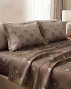 A bed dressed in a brown bedding set with a matching duvet cover and two pillowcases, featuring a wavy abstract pattern in taupe and white with thin olive-green piping, shown in a softly lit bedroom setting.