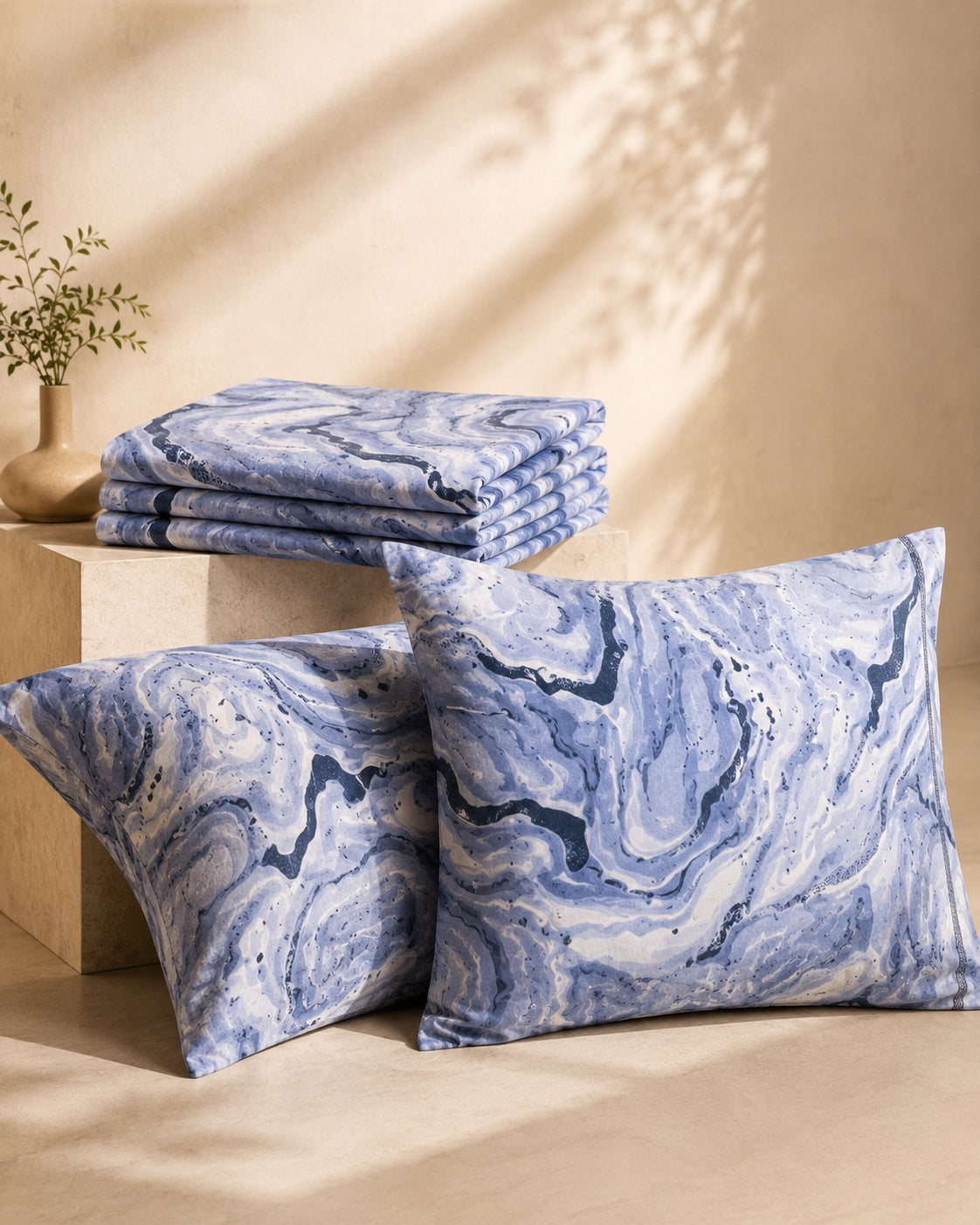 Three blue and white marble-patterned throw pillows and a matching folded textile stack, styled on beige blocks against a warm beige wall with soft leaf shadows.