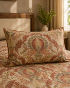 A rectangular decorative throw pillow with an ornate floral tapestry pattern in rust, beige, and muted green, resting on a matching patterned bedspread in a softly lit bedroom.