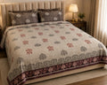 A beige quilted bed with a patterned duvet cover and matching pillows, styled in a warm, neutral-toned bedroom setting with a bedside lamp and curtains.