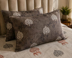 Two rectangular throw pillows with a dark brown textured fabric and a white tree pattern, styled on a bed with a matching beige duvet and a beige tufted headboard.