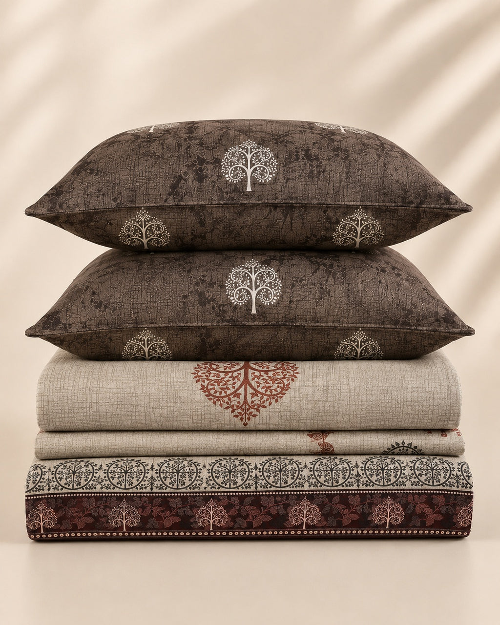 A stack of home textiles featuring two dark brown pillows with white tree motifs, a folded beige textile with a rust-red tree design, and a folded dark patterned sheet or blanket at the bottom, set against a warm beige background with soft leaf shadows.