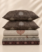 A stack of home textiles featuring two dark brown pillows with white tree motifs, a folded beige textile with a rust-red tree design, and a folded dark patterned sheet or blanket at the bottom, set against a warm beige background with soft leaf shadows.