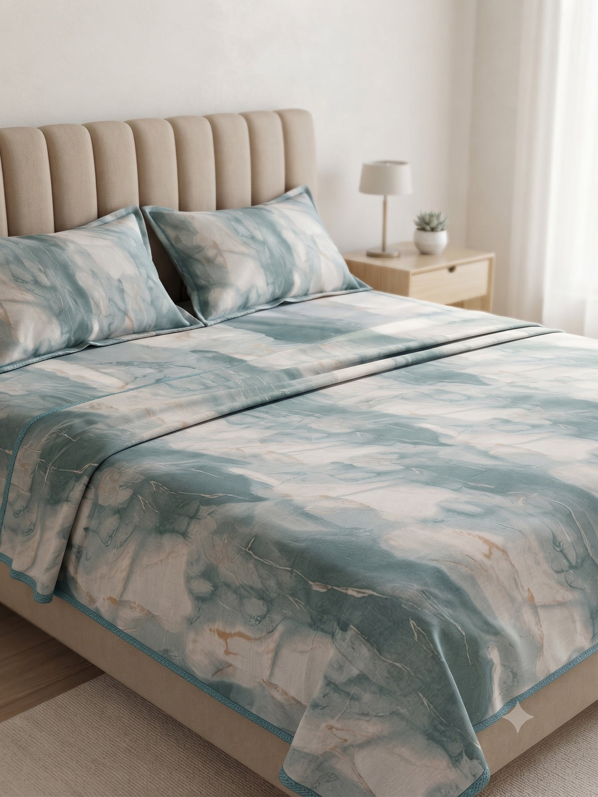 A neatly made bed in a softly lit bedroom, covered with a watercolor-style blanket and matching pillow shams in muted teal, blue, white, and beige tones. A beige upholstered headboard, small nightstand with a lamp, and sheer curtains are visible in the room.