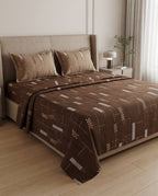 A bed dressed in a brown patterned quilt and matching pillow shams, styled in a modern bedroom with a beige upholstered headboard, bedside lamp, and light wood flooring.