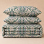 A stack of three matching upholstered home textiles with an ornate damask-style floral pattern in muted green and lavender on a warm beige background, photographed against a plain beige backdrop.