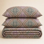 Three stacked pieces of patterned bedding, including two pillows and a folded blanket or comforter, in a dusty purple and sage green geometric design on a beige background.