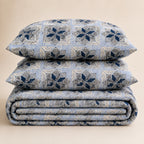 A stack of three bedding pieces, including two pillows and a folded blanket, featuring a blue and beige geometric damask pattern on a warm beige background.