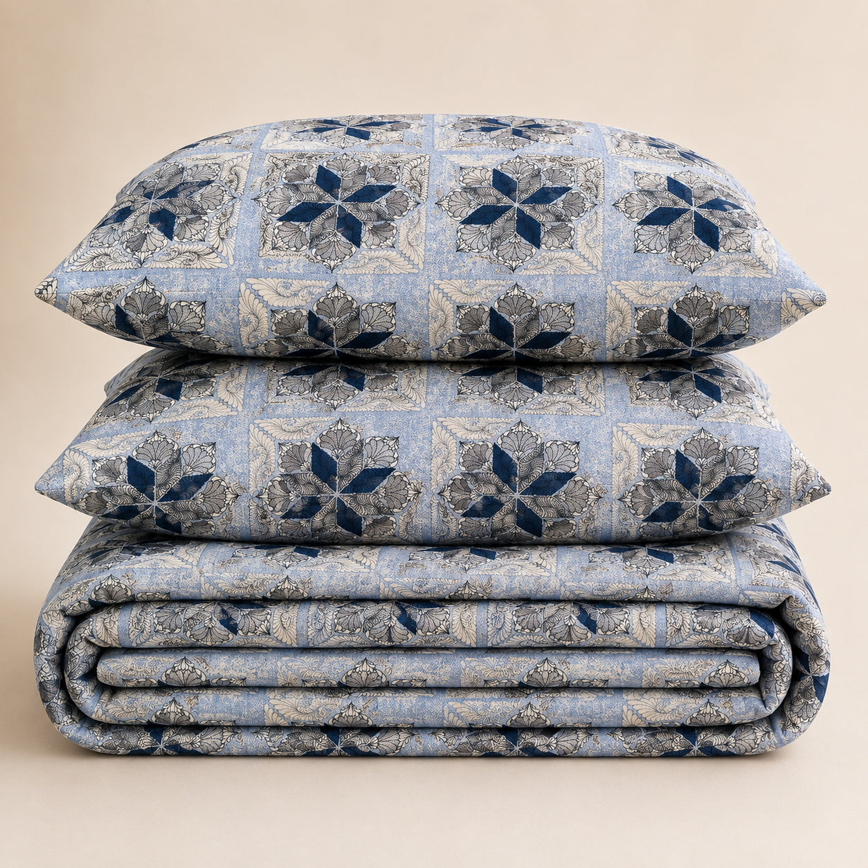 A stack of three bedding pieces, including two pillows and a folded blanket, featuring a blue and beige geometric damask pattern on a warm beige background.