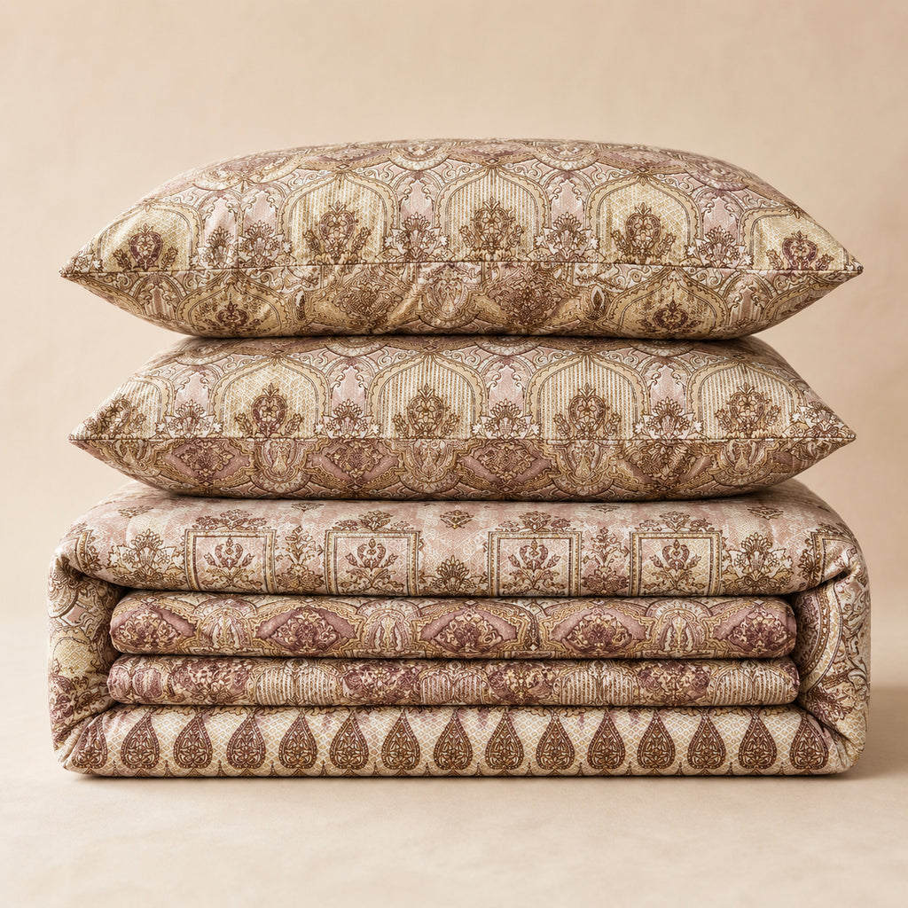 A stack of three matching decorative pillows and folded bedding pieces with an ornate damask-style pattern in warm beige, taupe, and brown tones, photographed against a plain beige studio background.