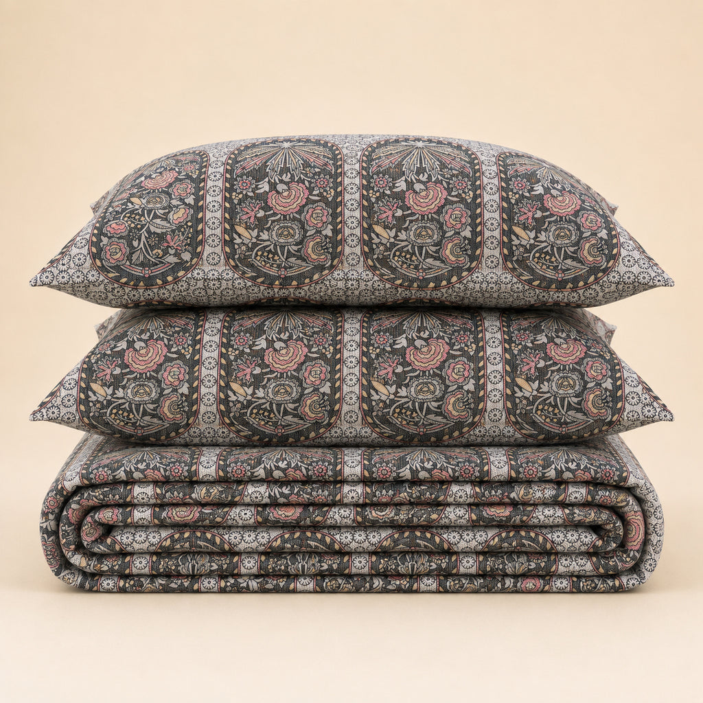 A stack of three patterned bedding pieces, including two pillows and a folded quilt or comforter, featuring a detailed hand-block print in black, gray, pink, and beige on a solid light beige background.