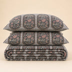 A stack of three patterned bedding pieces, including two pillows and a folded quilt or comforter, featuring a detailed hand-block print in black, gray, pink, and beige on a solid light beige background.