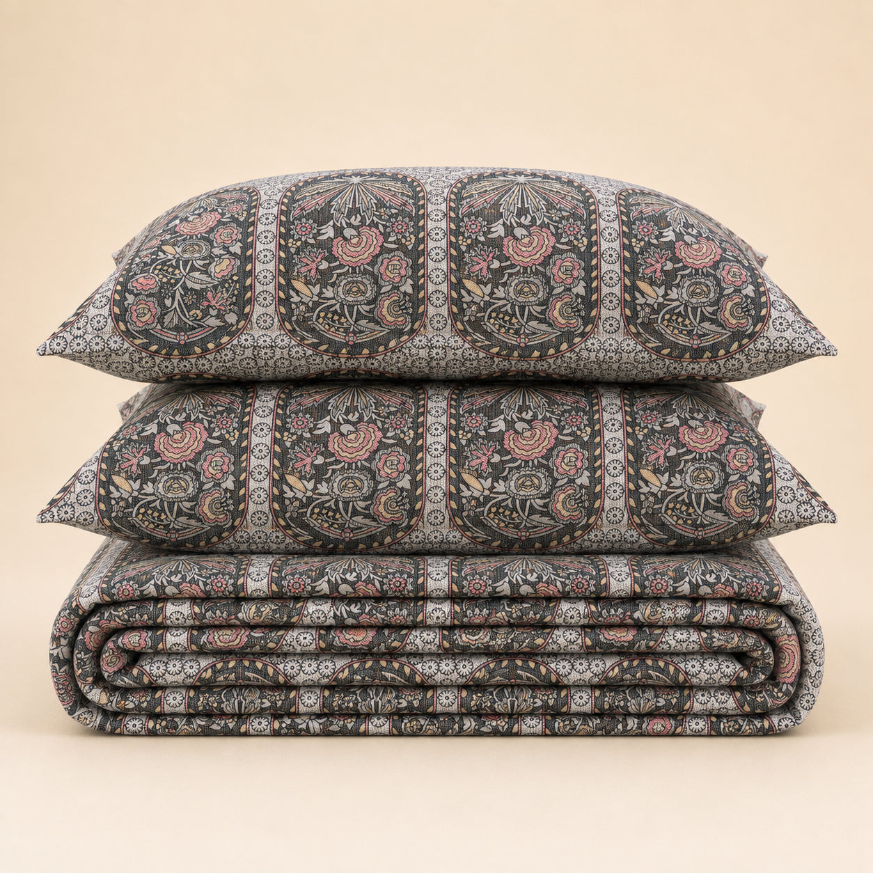 A stack of three patterned bedding pieces, including two pillows and a folded quilt or comforter, featuring a detailed hand-block print in black, gray, pink, and beige on a solid light beige background.