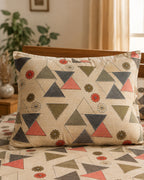 A decorative rectangular throw pillow with a geometric pattern of triangles and small floral motifs in muted red, olive green, navy, and cream, resting on a bed with matching pillowcases and a matching bedspread.